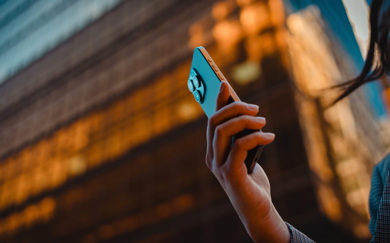 A hand holding a phone with a teal screen, a warm-lit building out of focus behind it.