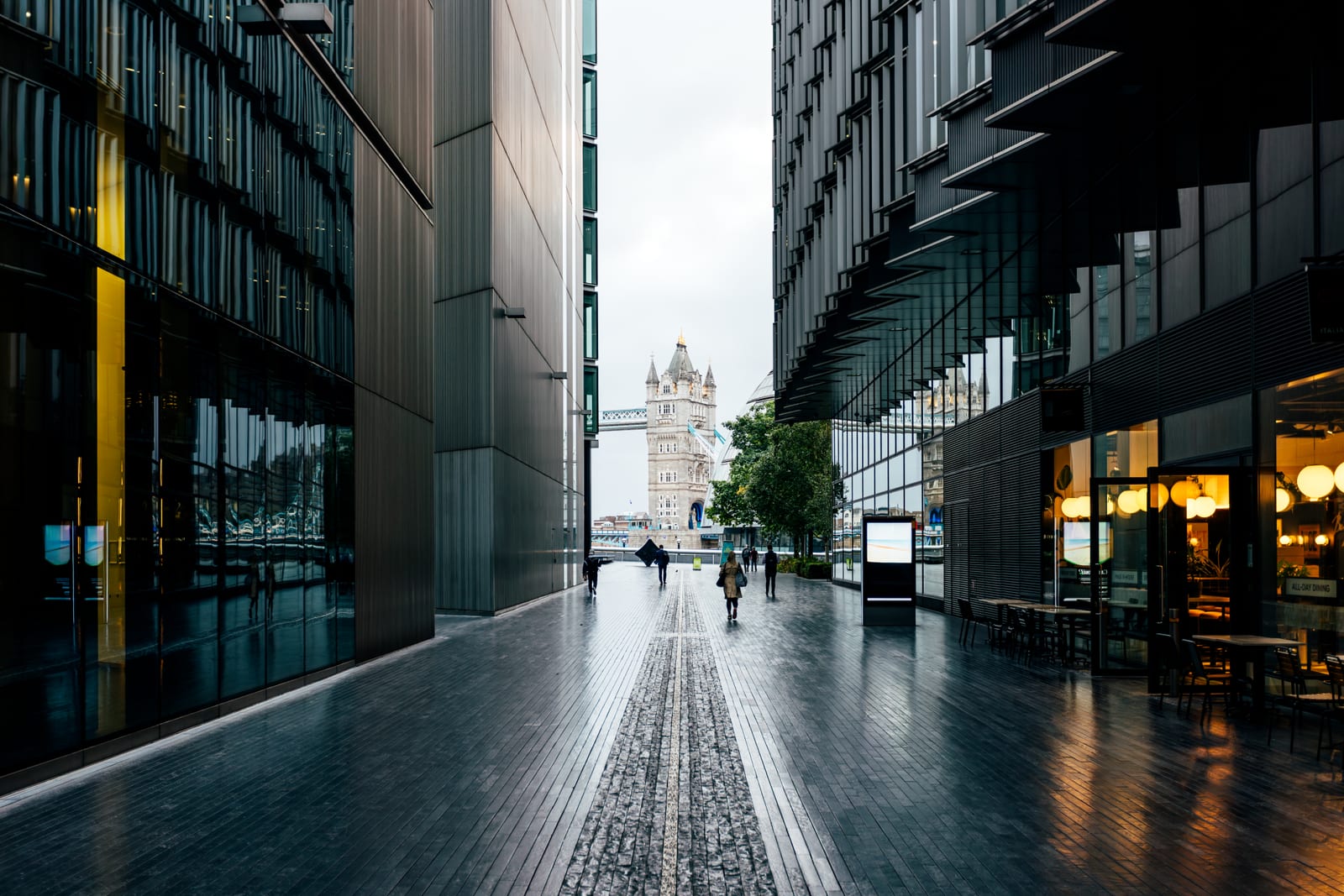 A wet city street between glass buildings at dusk, a bridge tower in the distance.